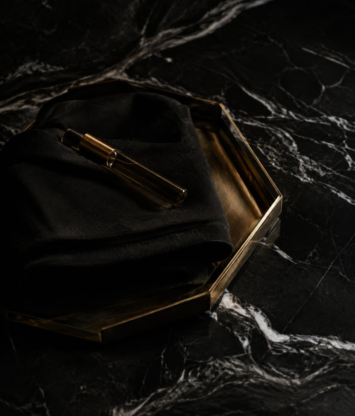 Gold vial on folded black linen in a brass tray on marble